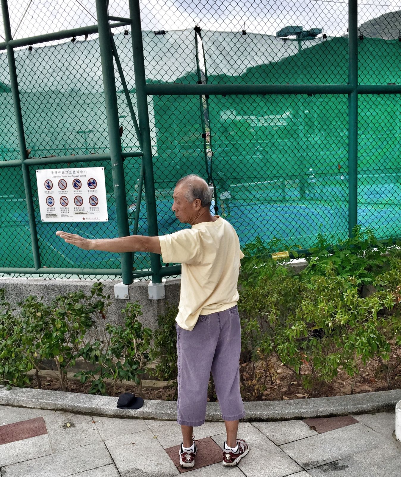Älterer Mann in hellem T-Shirt und lila Hose übt Tai Chi vor dem grünen Zaun des Aberdeen Tennis and Squash Centre in Hongkong. Er streckt einen Arm mit flacher Hand zur Seite, seine Mütze liegt auf dem Mäuerchen.