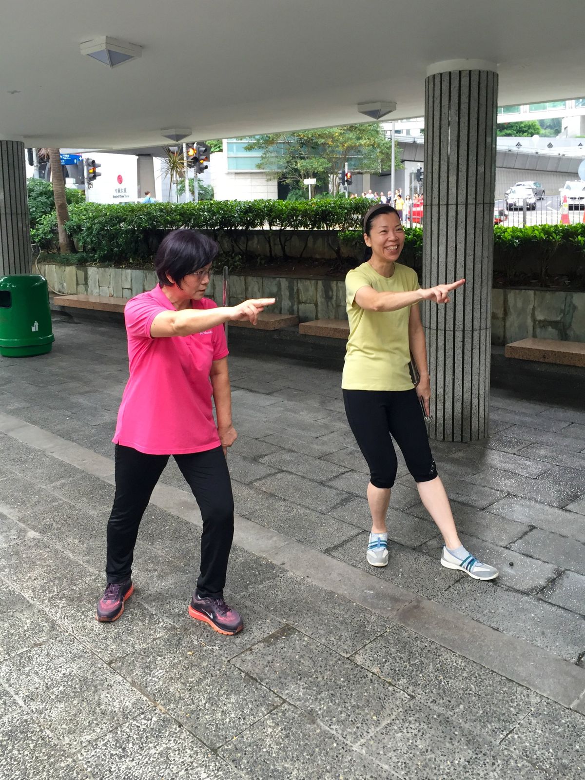 Zwei Frauen in Sportkleidung unter einem Vordach mit Granitsäulen in Central, Hongkong. Beide strecken einen Arm aus und zeigen mit gestreckten Fingern in dieselbe Richtung. Im Hintergrund Hecke, Straße und ein Schild des Bank of China Tower.