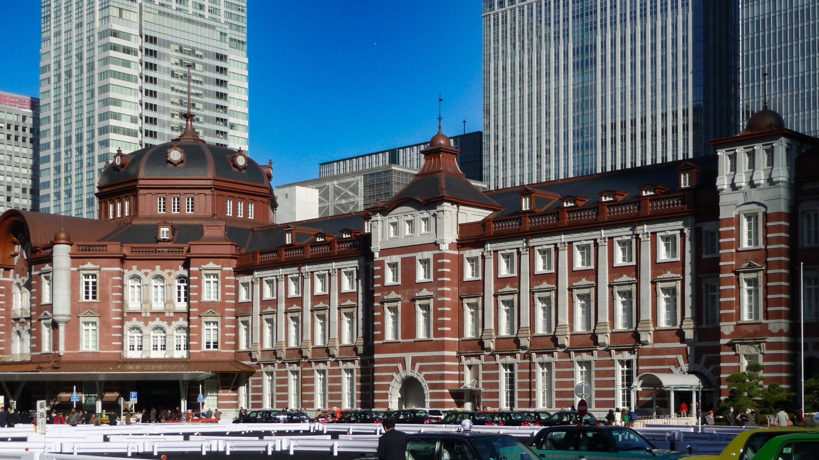 Rotes Backsteingebäude des Bahnhofs Tokyo auf der Marunouchi-Seite mit Kuppel, davor Taxis, dahinter Hochhäuser