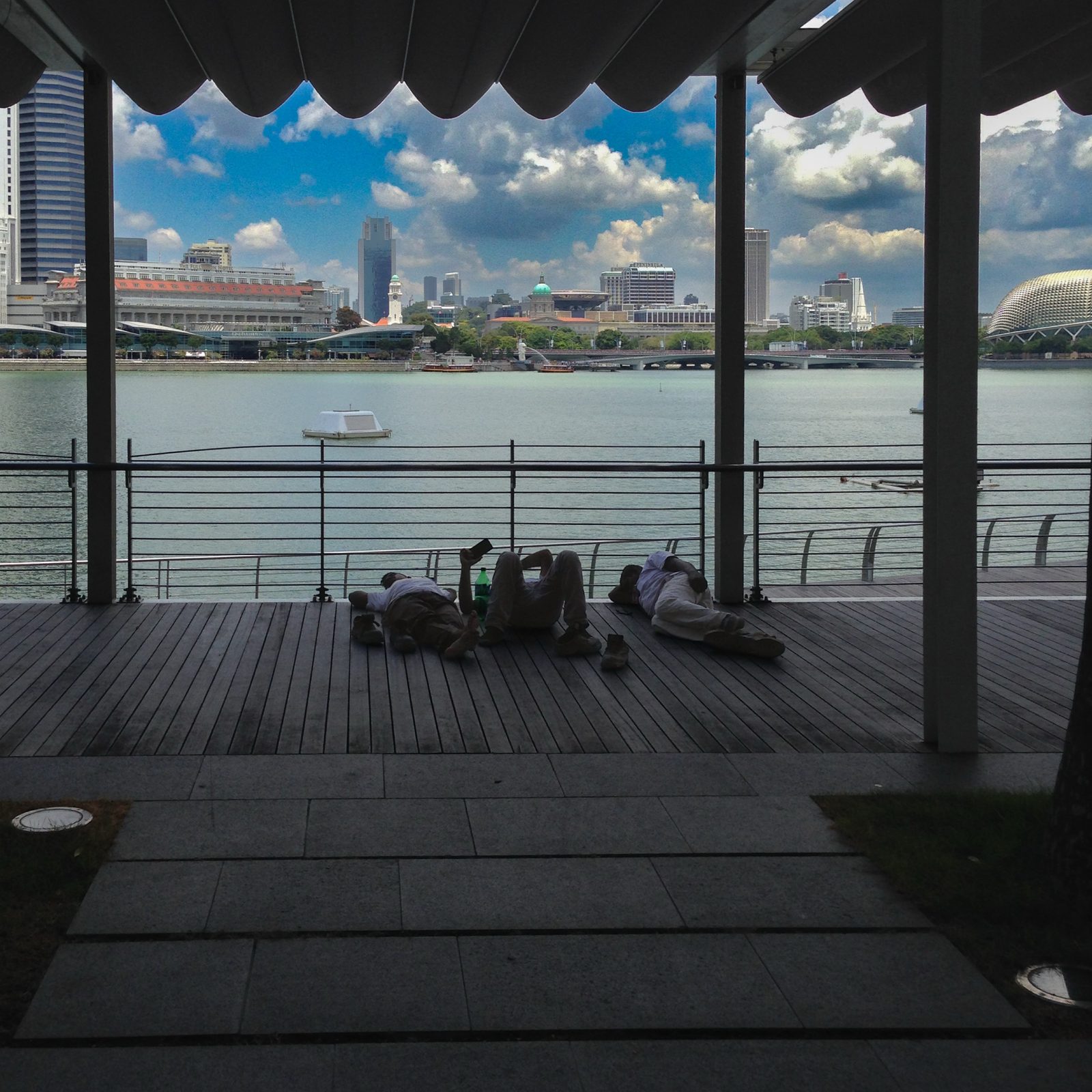 Drei Männer machen Mittagsschlaf auf einem Holzdeck an der Marina Bay, dahinter die Skyline