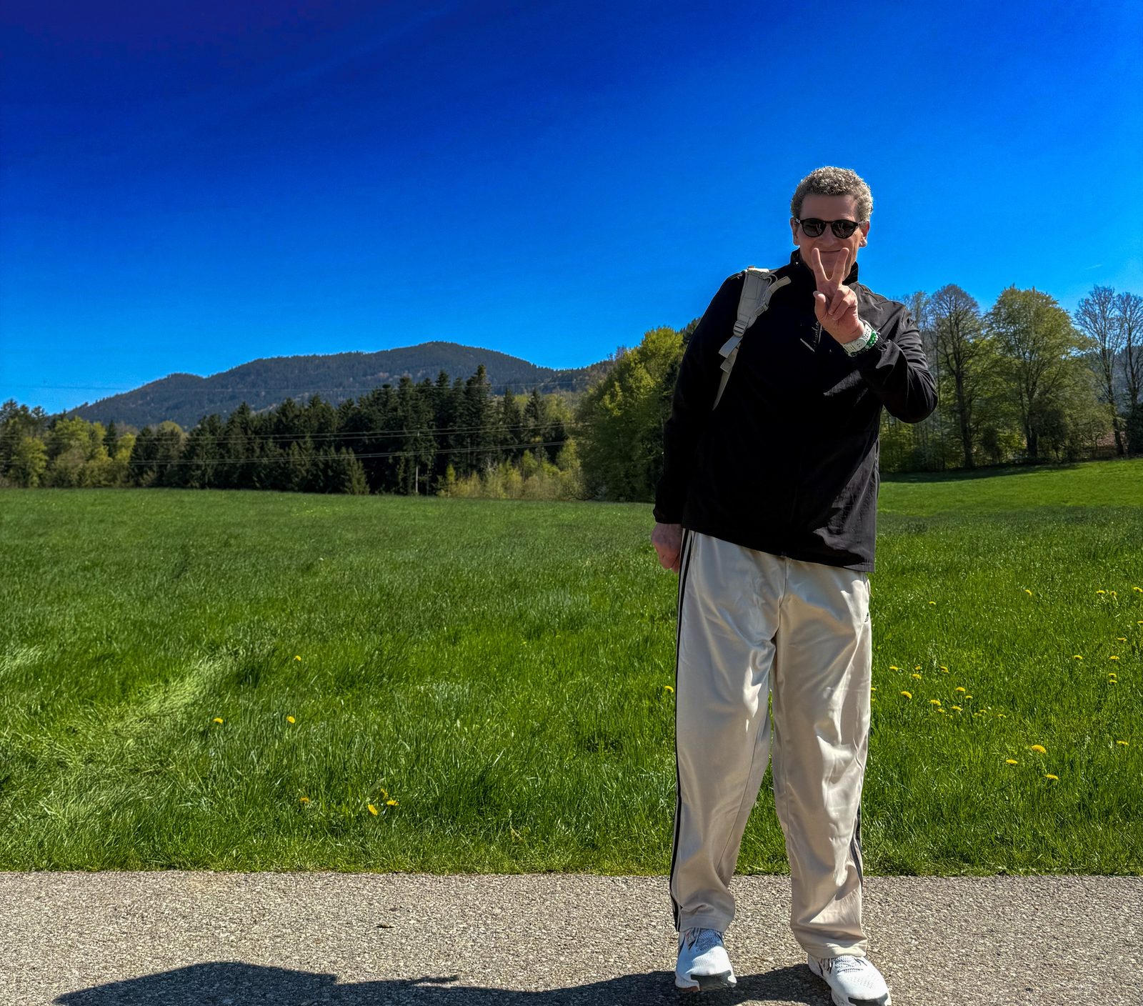 Torsten Fink mit Peace-Zeichen auf einem Wanderweg vor grünen Wiesen und Voralpen