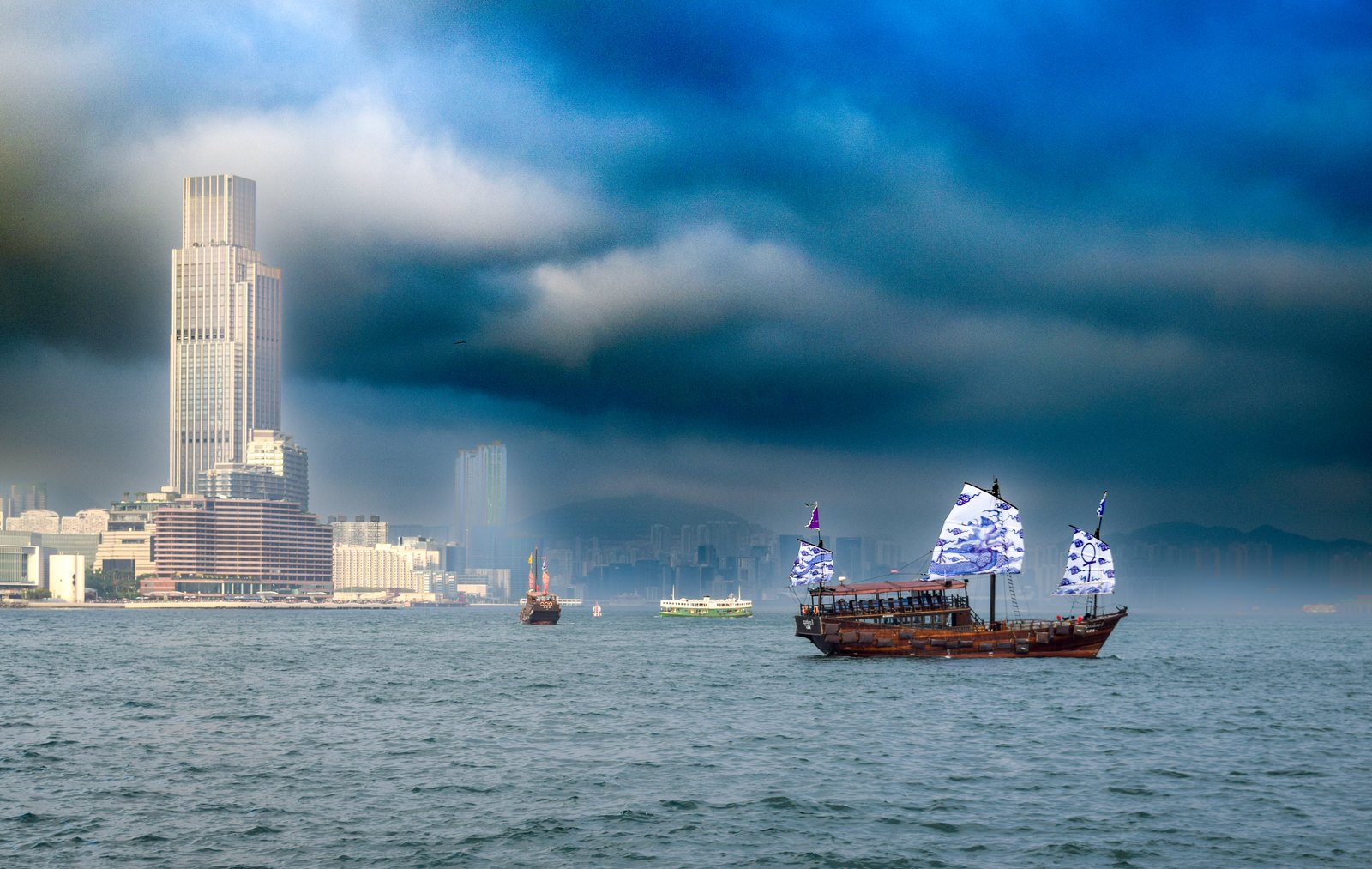 Traditionelle chinesische Dschunke mit blau bedruckten Segeln vor der Skyline von Tsim Sha Tsui unter dramatischem Wolkenhimmel