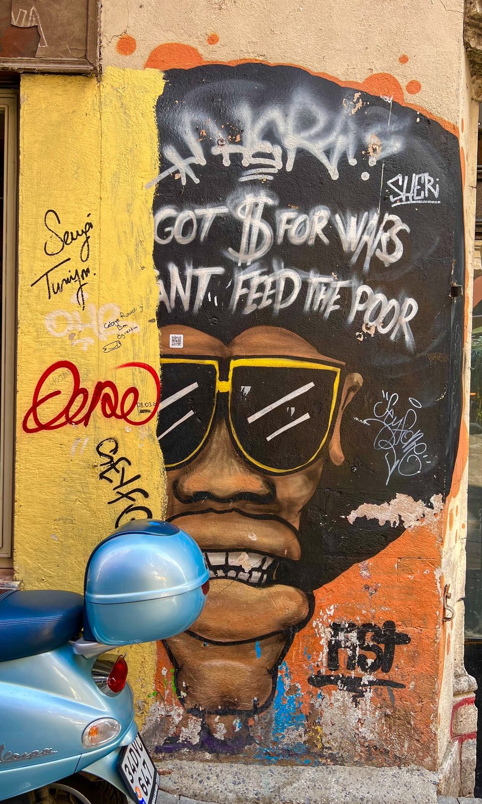 Großes Mural eines Gesichts mit Sonnenbrille und gesellschaftskritischem Schriftzug, davor eine hellblaue Vespa