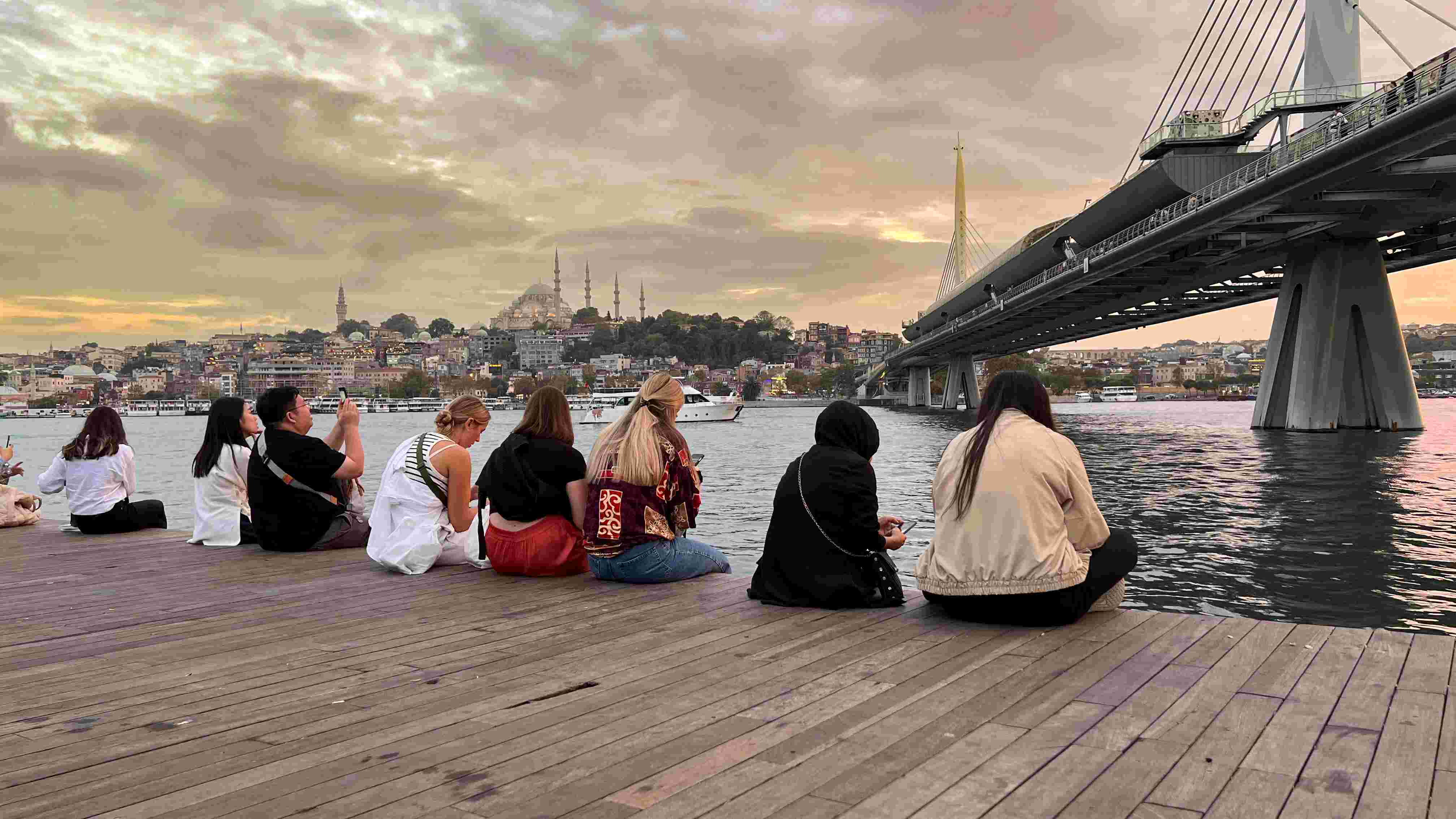 Menschen sitzen nebeneinander auf Holzplanken am Goldenen Horn und schauen aufs Wasser