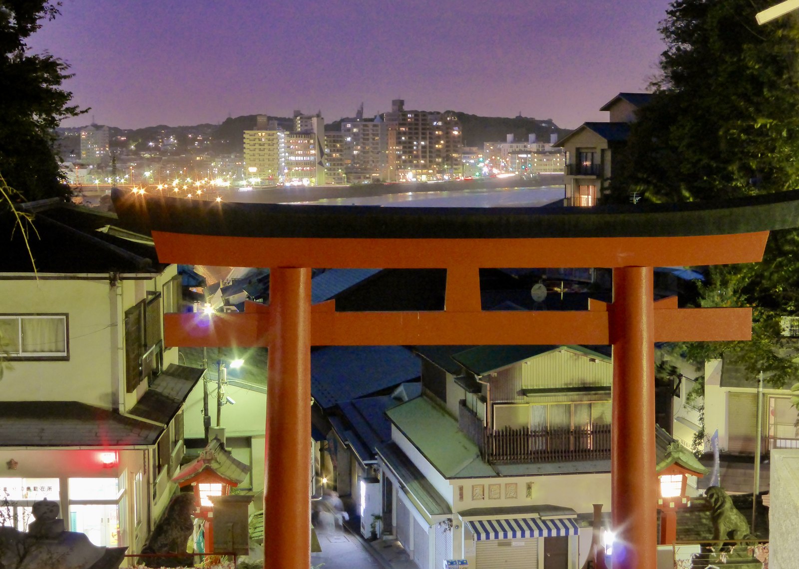 Rotes Torii des Nakatsu-no-miya-Schreins bei Nacht, Blick hindurch auf die Lichter Richtung Kamakura und die Brücke zum Festland