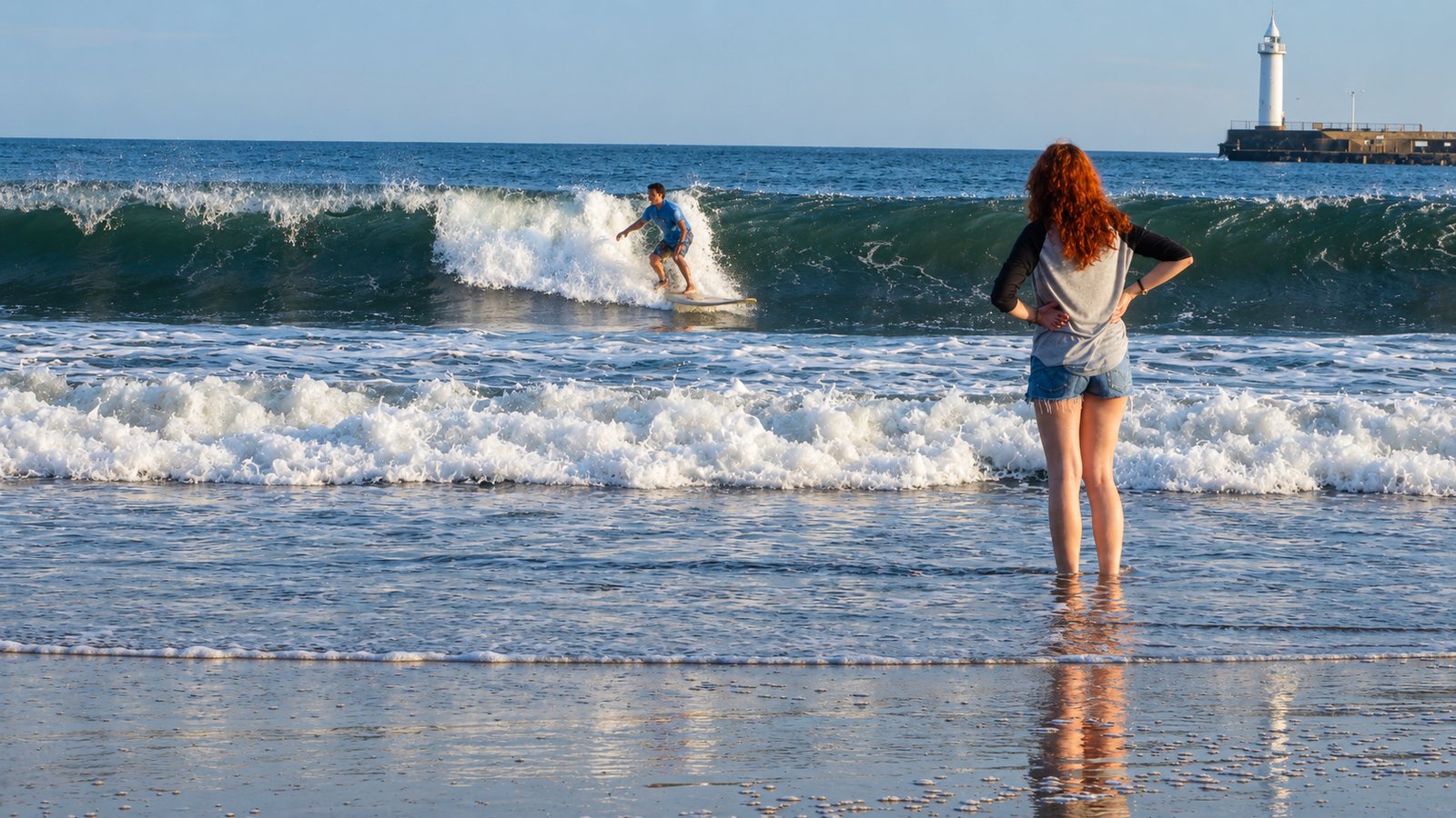 Laura, die Tochter von Torsten Fink, steht mit roten Locken bis zu den Waden im Meer am Strand von Yuigahama in Kamakura und beobachtet einen Surfer auf einer brechenden Welle, rechts ein weißer Leuchtturm auf einer Mole
