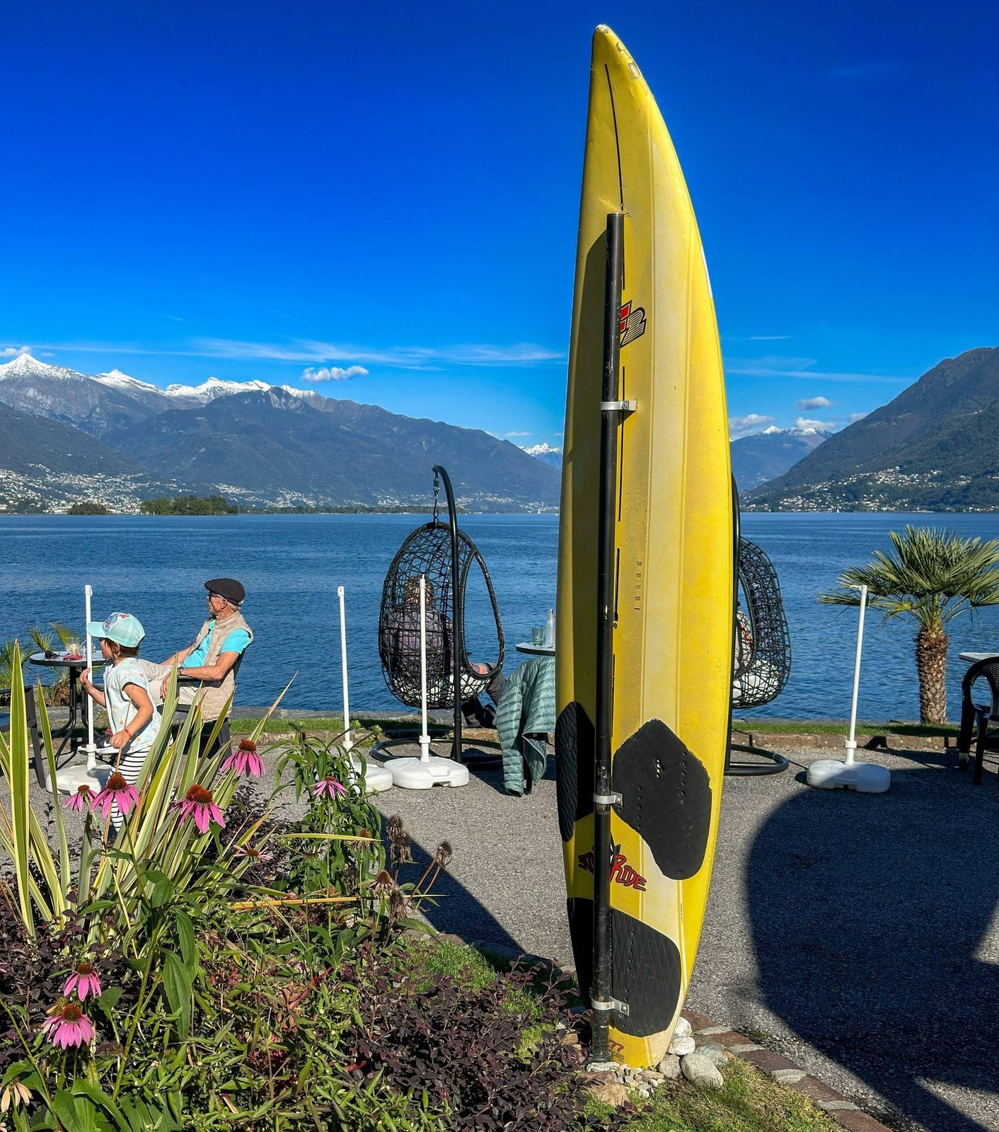 Gelbes Surfbrett und Hängesessel am Ufer des Lago Maggiore mit schneebedeckten Bergen