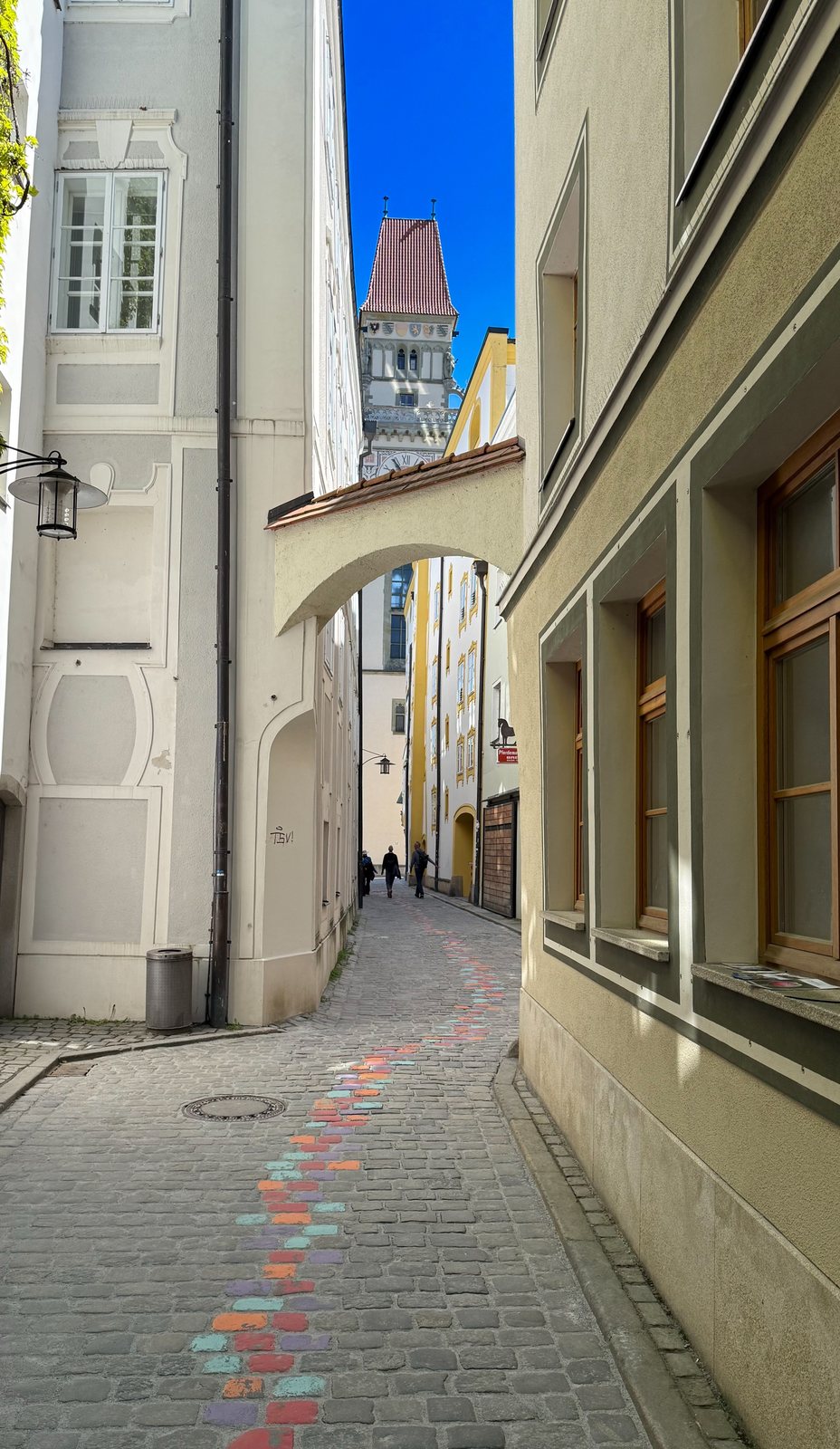 Schmale Gasse mit bunt bemaltem Pflaster und Blick durch einen Torbogen auf den Passauer Rathausturm