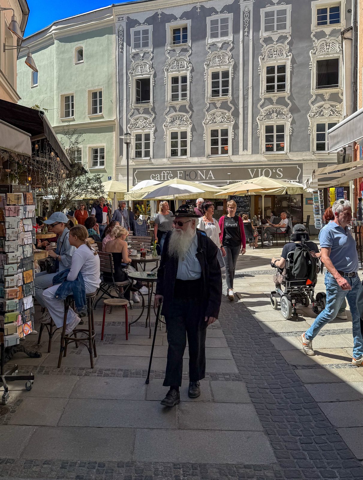 Belebter Passauer Marktplatz mit Stuckfassade, Straßencafés und einem älteren Herrn mit weißem Bart und Gehstock