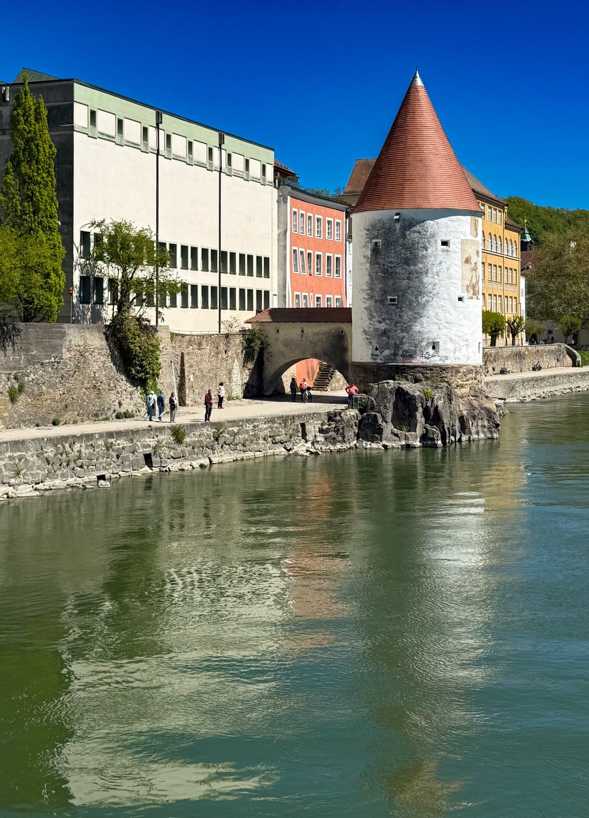 Runder Wehrturm mit rotem Kegeldach am Ufer der Ilz in Passau, davor bunte Altstadtfassaden