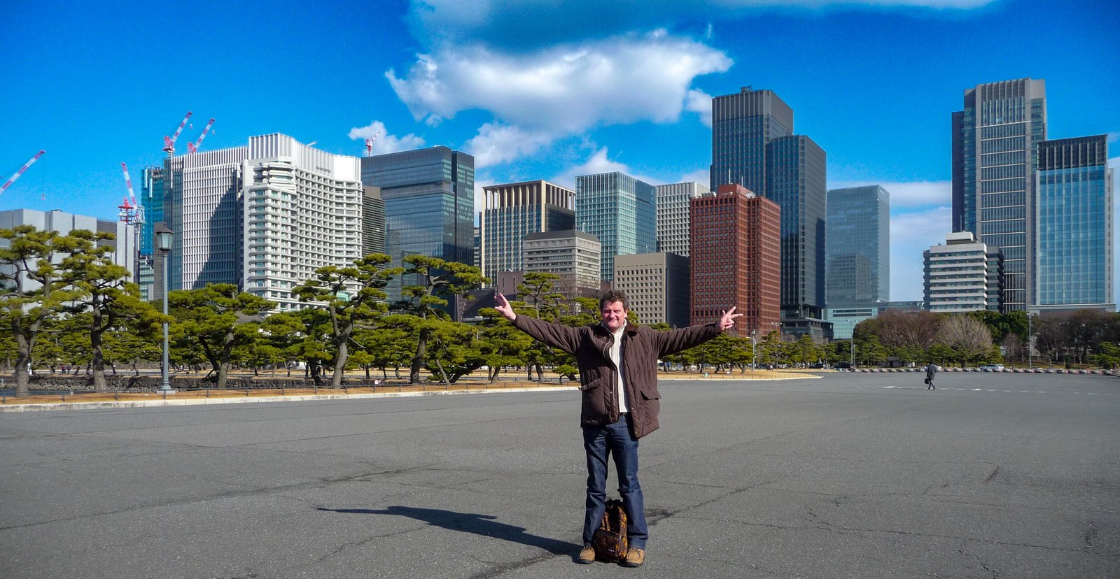 Torsten Fink mit ausgebreiteten Armen vor der Skyline des Finanzviertels Marunouchi, im Vordergrund Schwarzkiefern der Kōkyo-Gaien-Gartenanlage